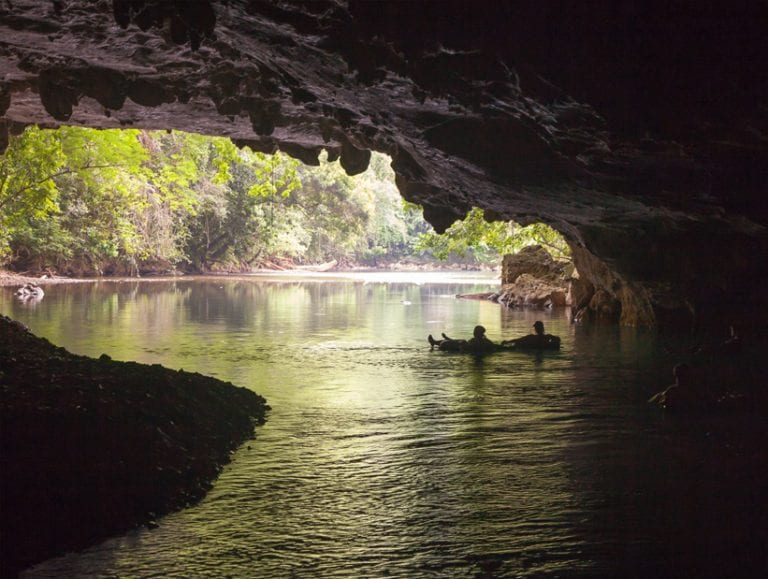 Two people cave tubing in Belize silhouetted inside a cave, floating toward the exit with forest view beyond