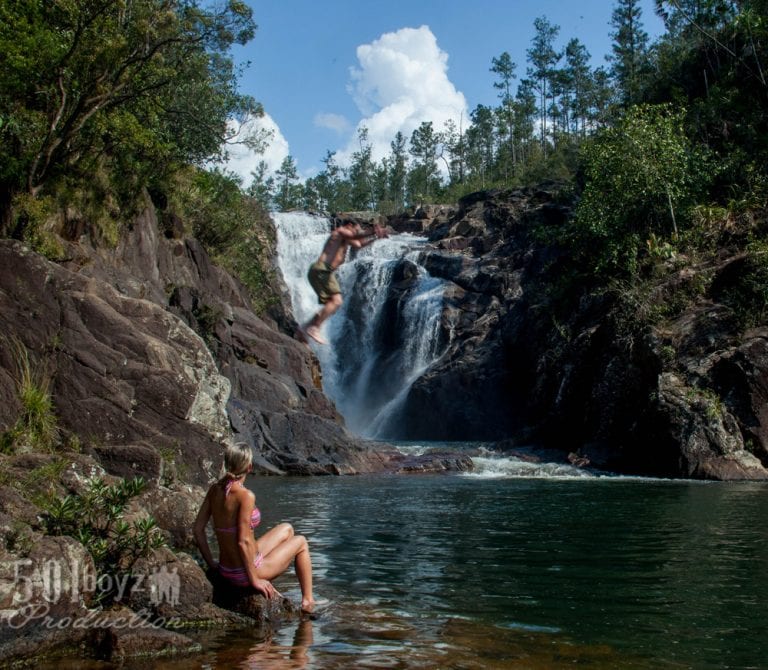 Couple swinging at Big Rock Falls in Belize with a man in the foreground and another man jumping off the rocks into the waterfall pool