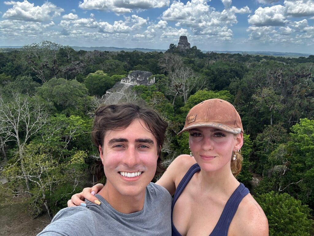Couple hugging and taking a selfie at Tikal in June 2025, with jungle canopy and Maya temple in the background
